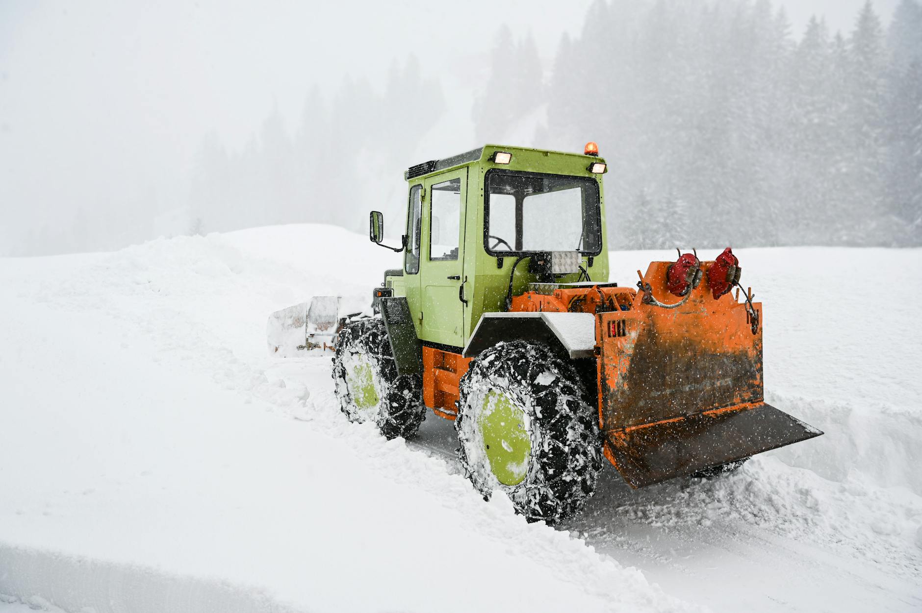 Vibrant bulldozer clears snow during a winter storm in a forested area.