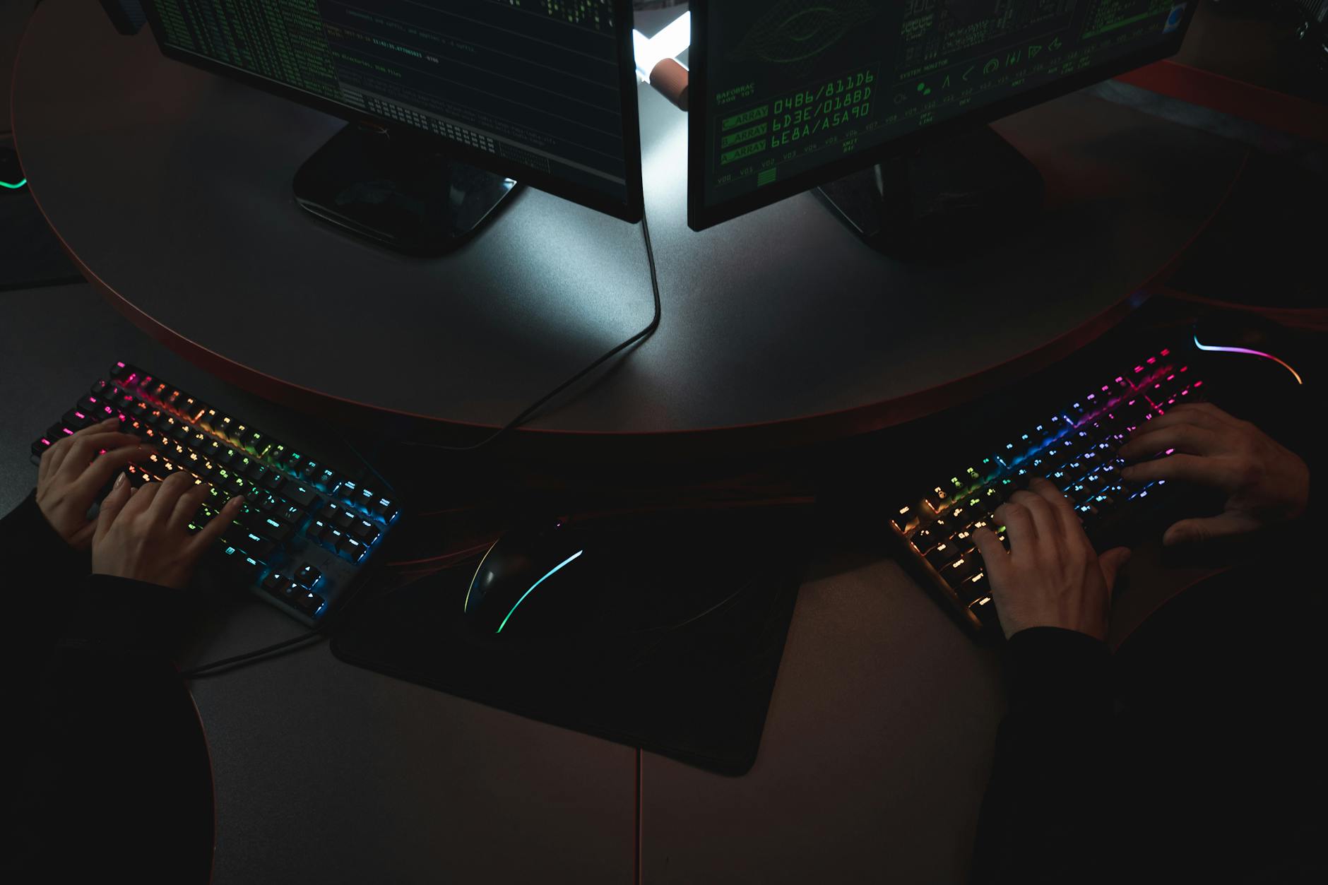 Two people typing on RGB keyboards with code on screens, indicating a cybersecurity environment.