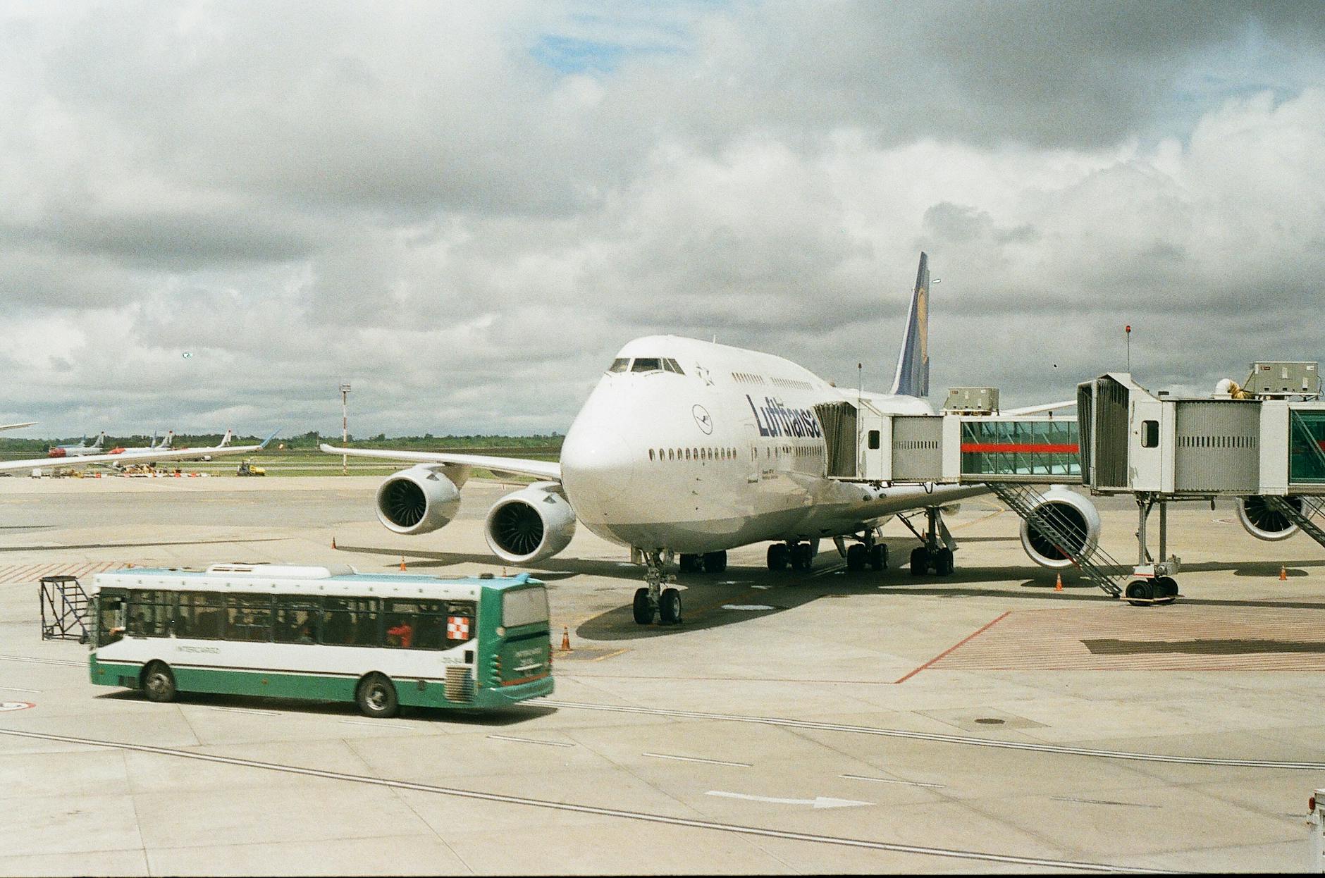 A large airplane docked at an airport gate with a shuttle bus nearby under cloudy skies.